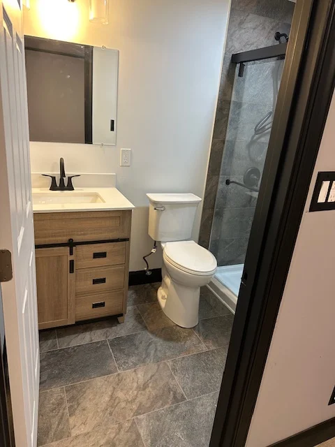 Painted bathroom with white walls, wood vanity, white toilet, and gray tile shower with black frame